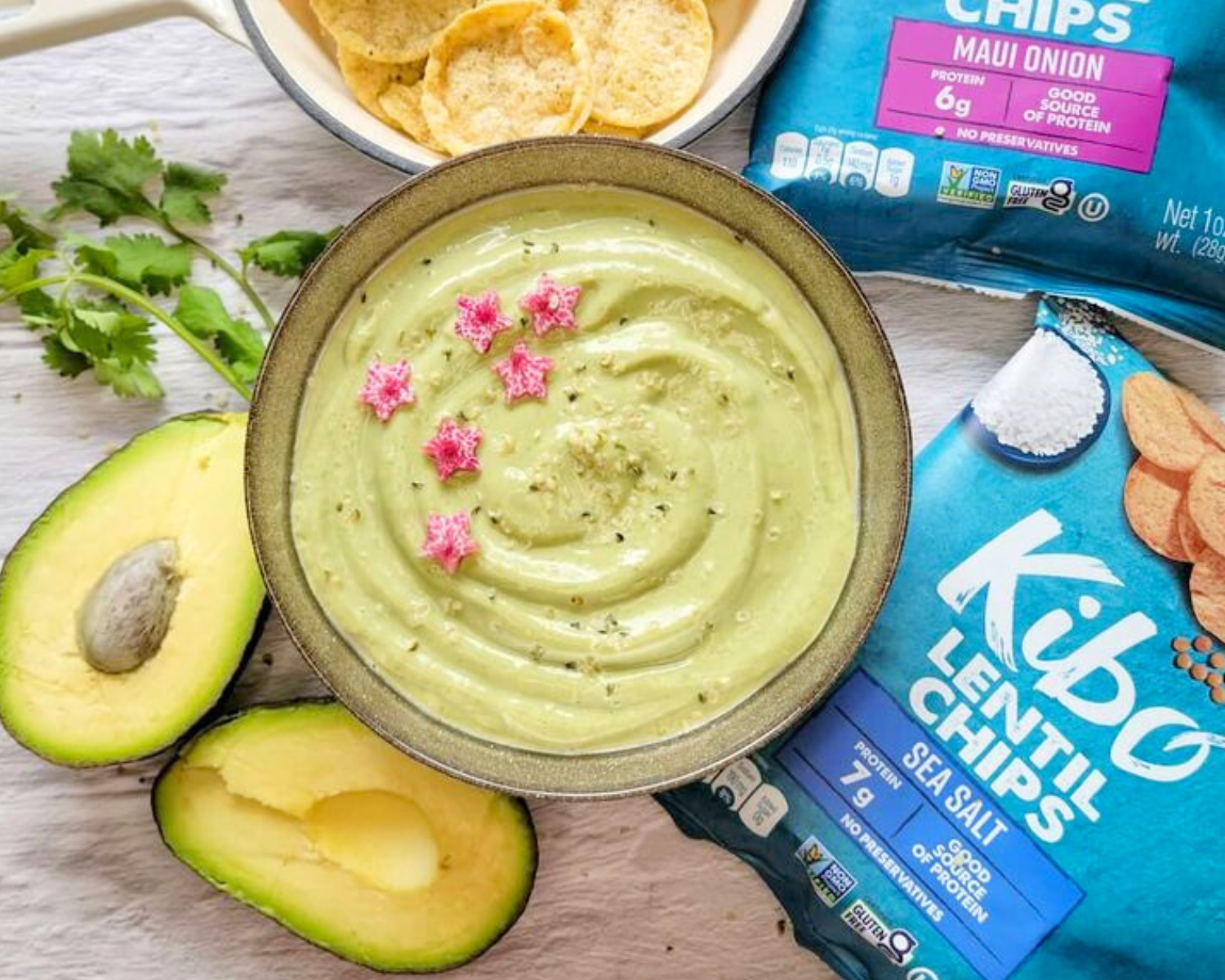 Green avocado dip in a bowl beside avocados and bags of Kibo Lentil Chips